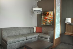 A modern hotel room with a grey sectional sofa, a wooden side table, a floor lamp, and a decorative wall panel.