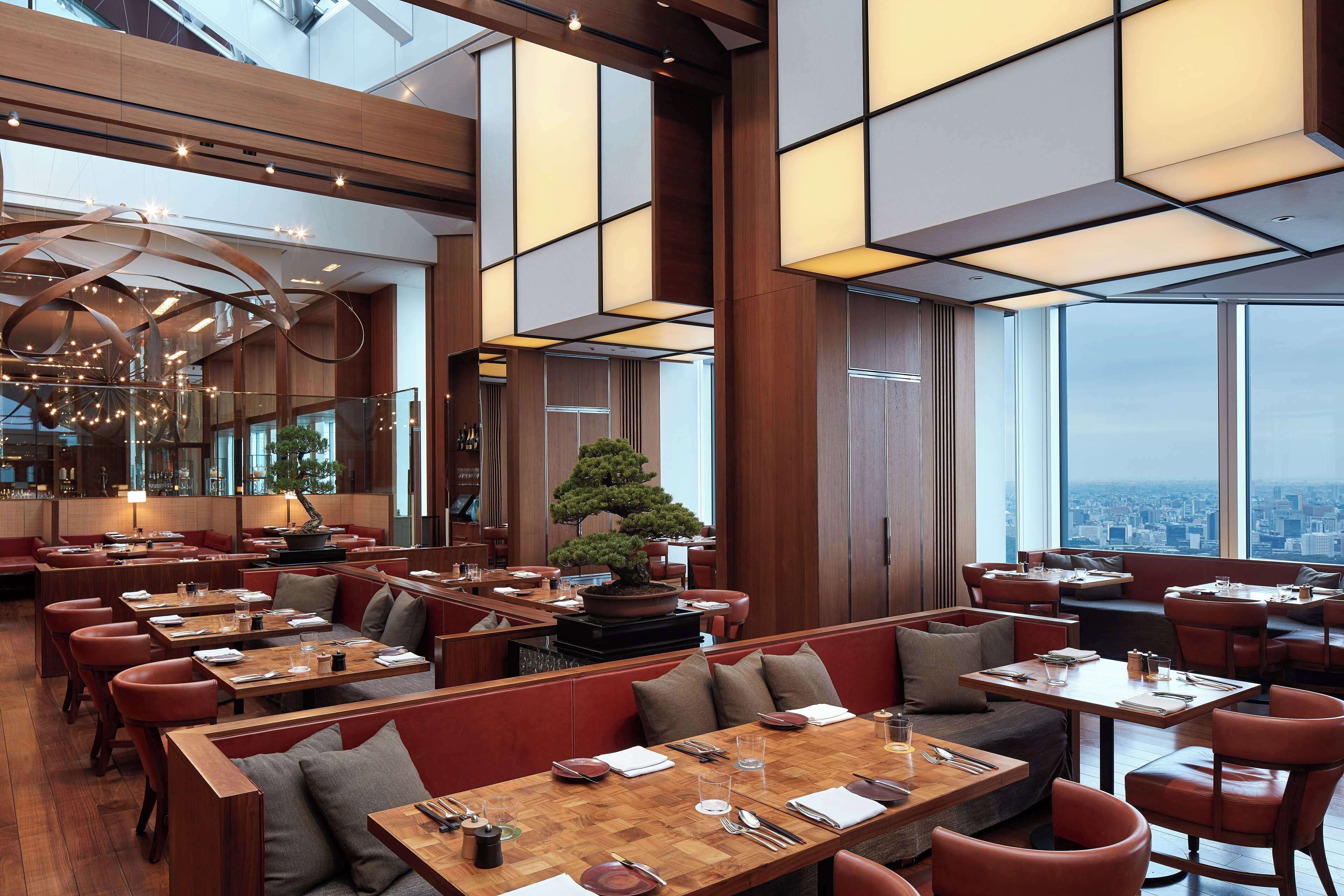 A modern restaurant with high ceilings, wooden paneling, and a city view. Tables are set with cutlery and glasses, and there are red leather booths.