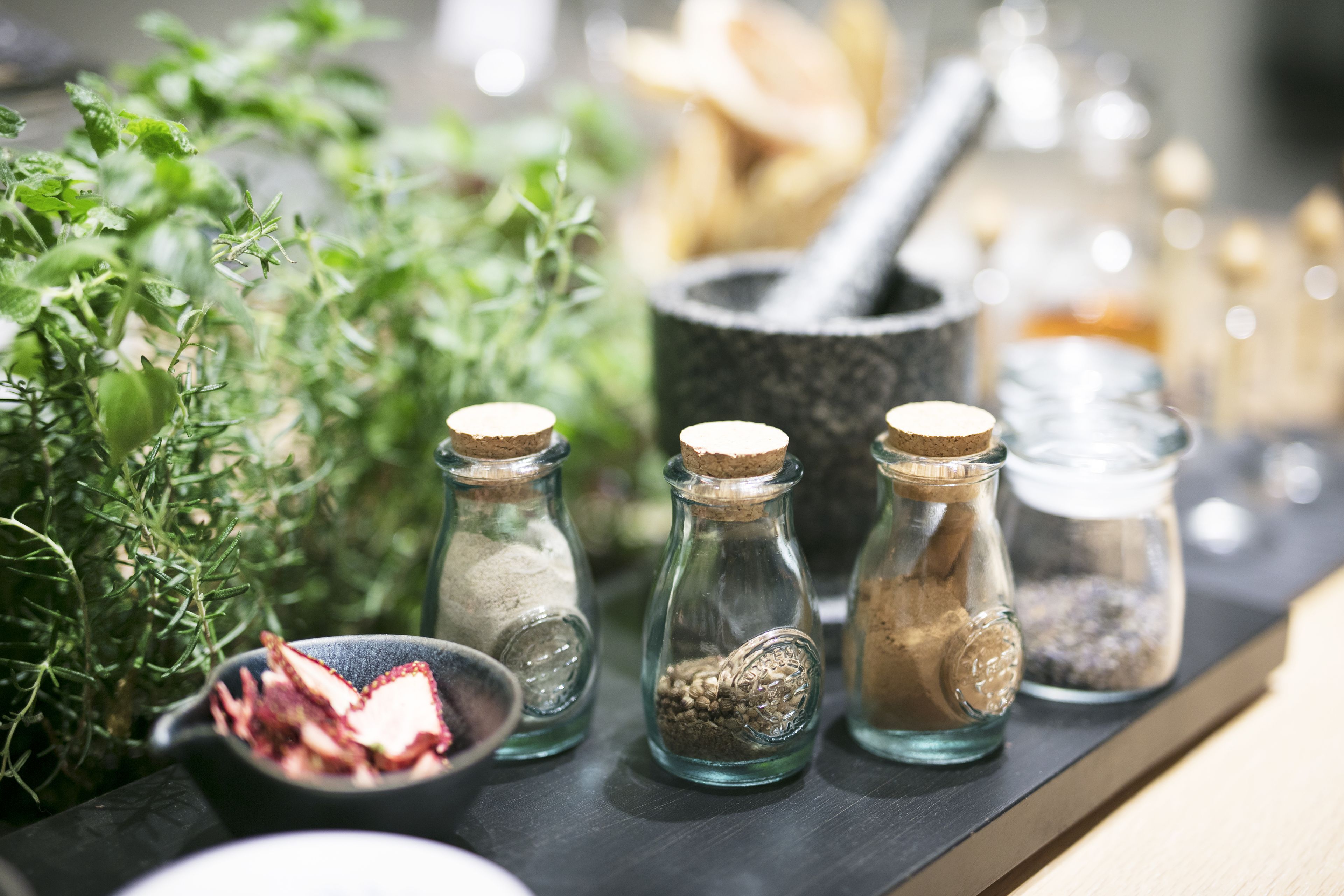 Des bocaux en verre avec des couvercles en liège contenant différentes épices et herbes aromatiques, un mortier et un pilon, et un bol rempli de piments rouges séchés, le tout posé sur une surface en bois.