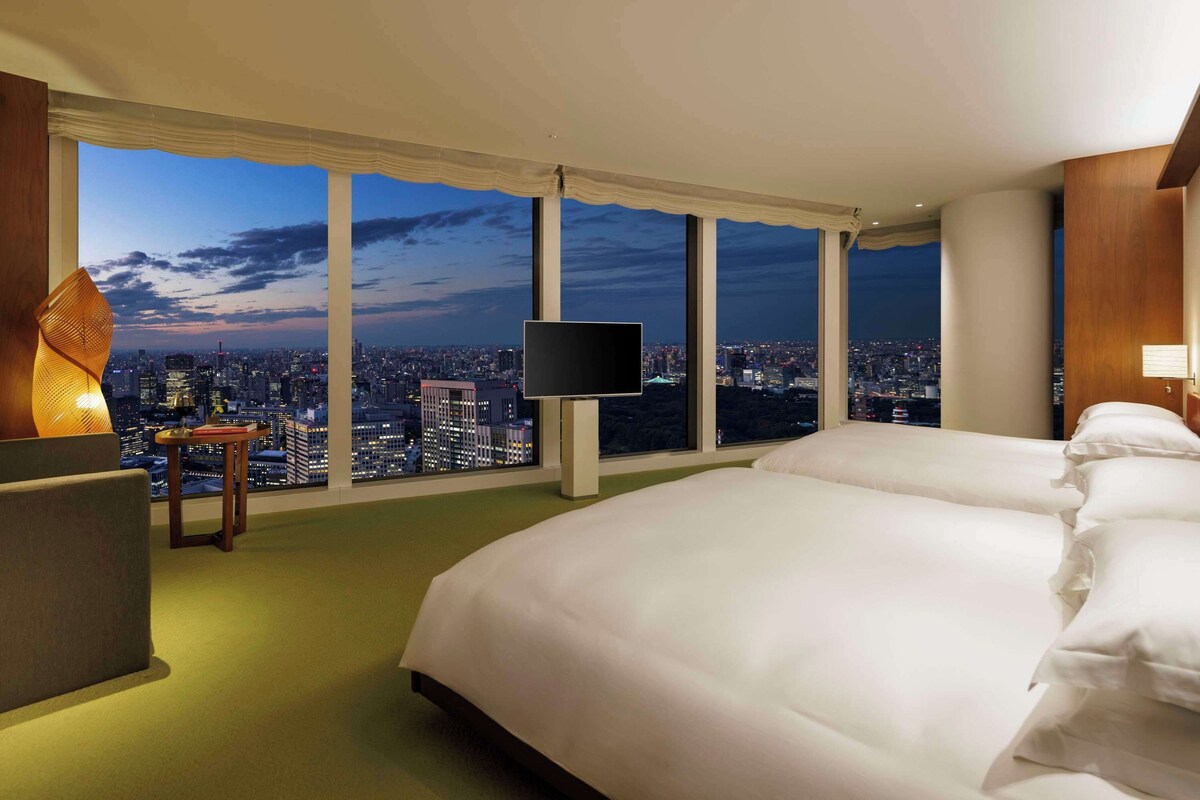 A hotel room with a large bed, a TV, a desk, and a view of a cityscape at night.