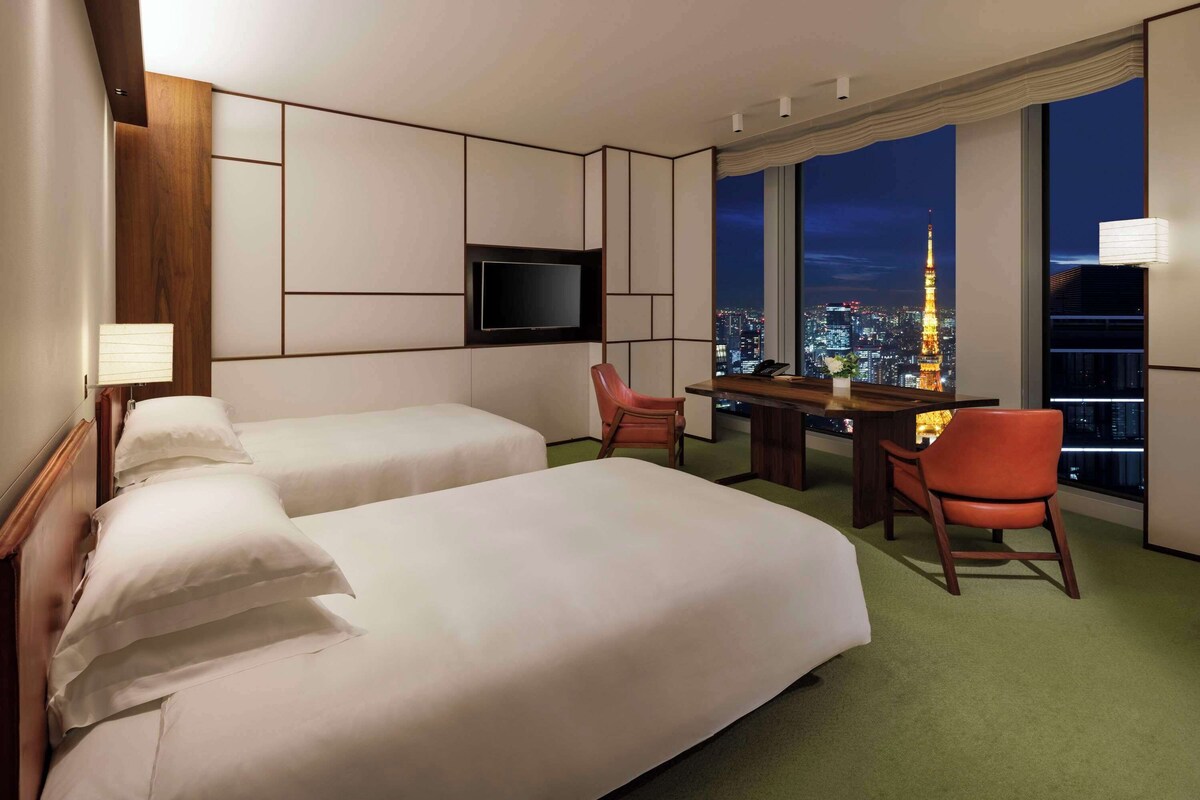 A hotel room with two beds, a desk, a chair, a TV, and a view of a cityscape at night.