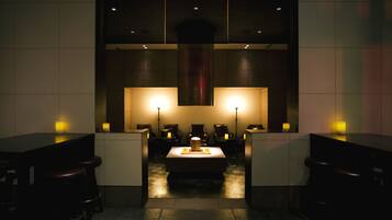 A dimly lit bar area with a central table and multiple stools, surrounded by walls with recessed lighting.