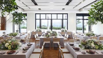 A spacious dining area with multiple tables set for a formal event, large windows offering a city view, and indoor plants adding greenery.