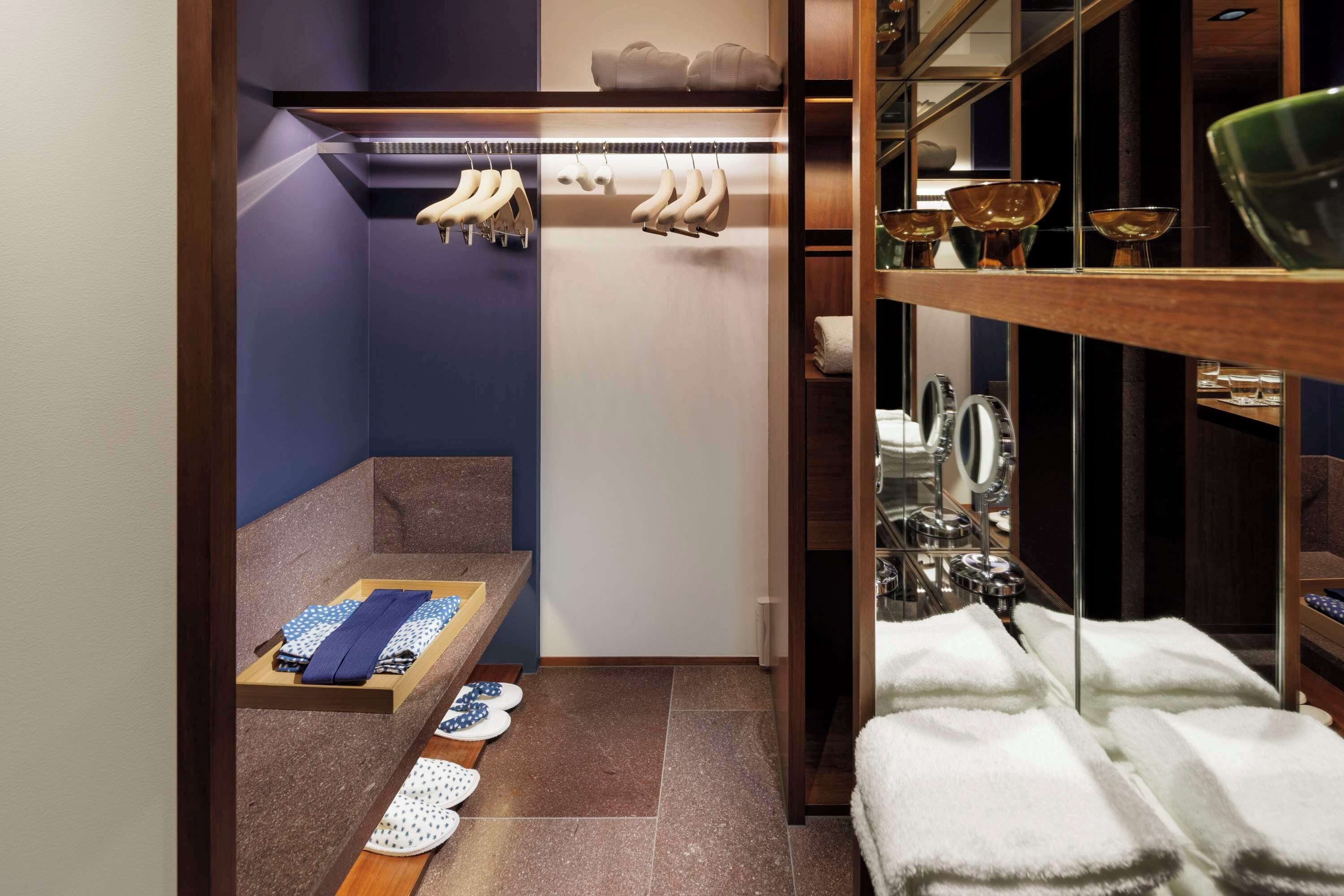 A compact hotel room with a built-in closet, a vanity with a mirror, and neatly arranged towels and shoes.