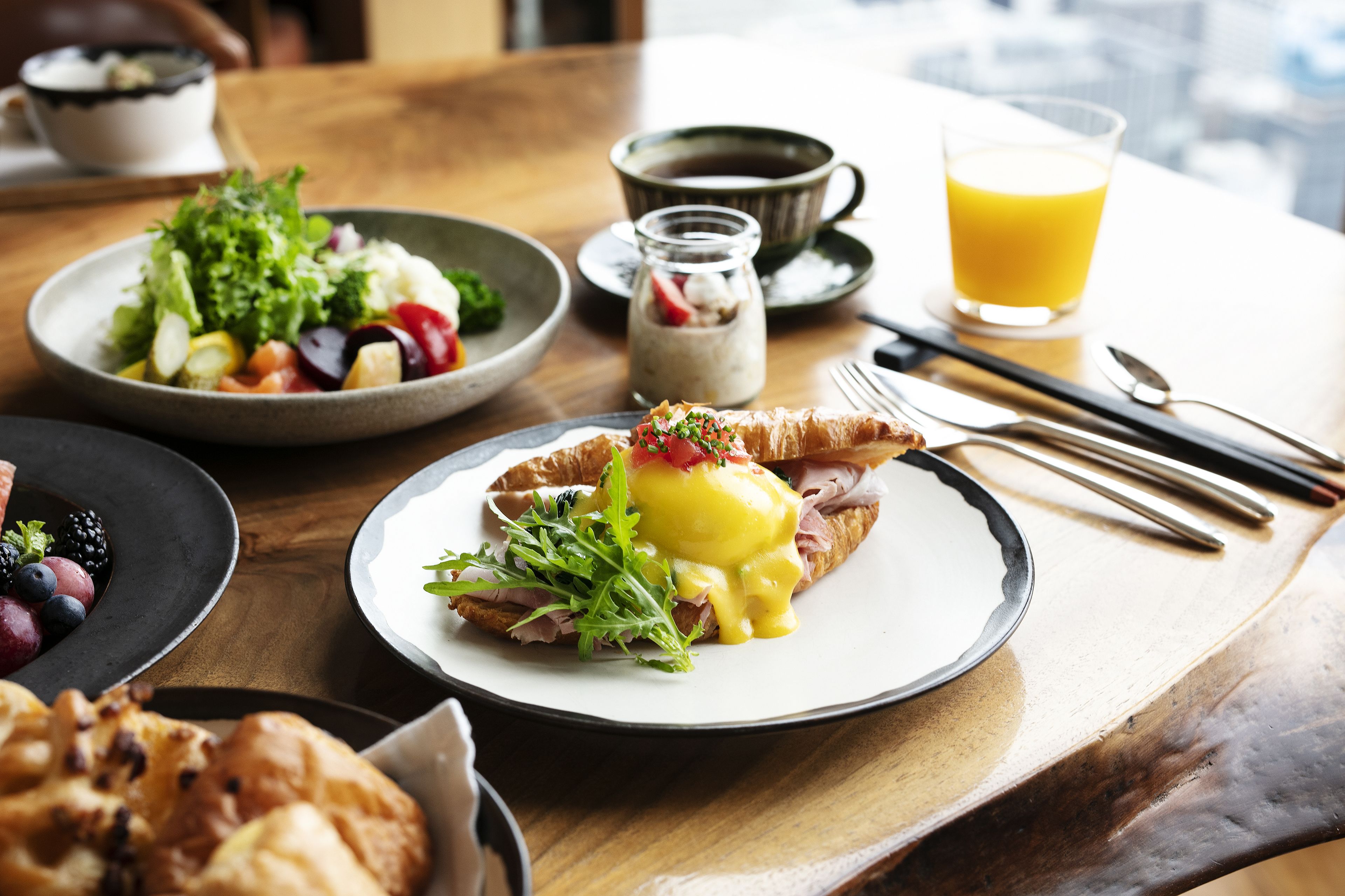 A breakfast table with a plate of eggs benedict, a bowl of fruit salad, a cup of coffee, and a glass of orange juice.