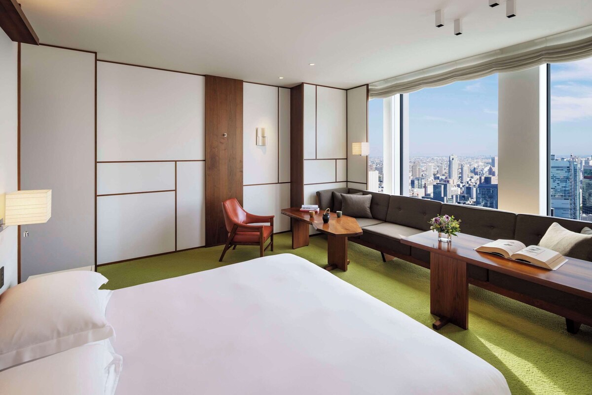 A modern hotel room with a large bed, a sofa, a chair, and a small table. There is a window with a city view.