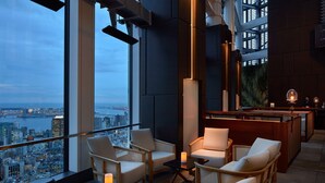 Een moderne lounge met uitzicht over de hoge stadspanden, comfortabele zitplaatsen en een bar.