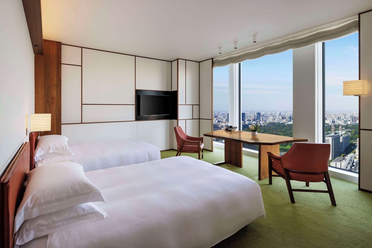 A hotel room with two beds, a desk, a chair, a TV, and a large window offering a city view.