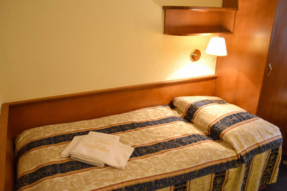 A neatly made bed with a patterned bedspread and pillows, a folded towel, and a wall-mounted lamp.