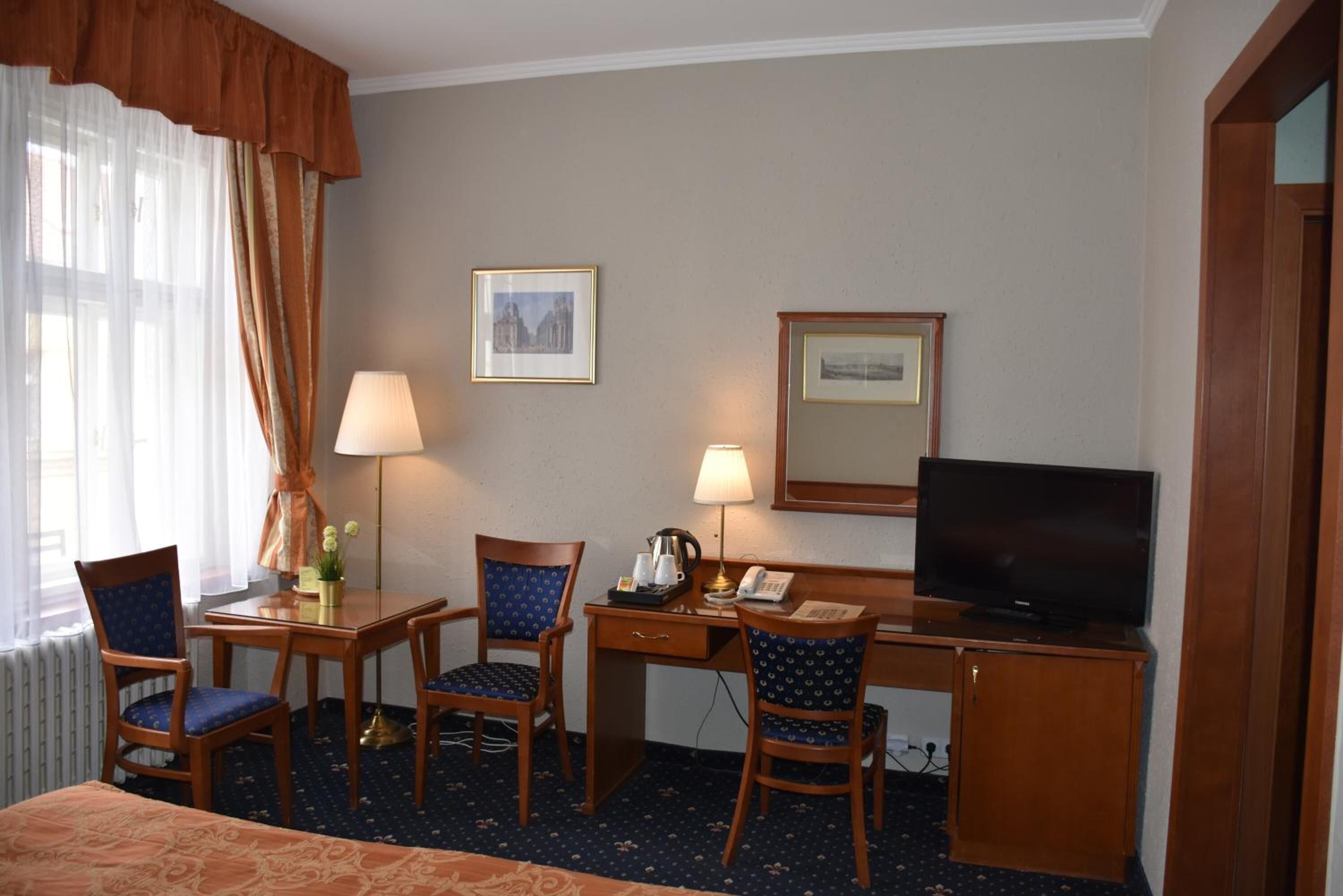A hotel room with a bed, two chairs, a desk with a lamp, a television, and framed pictures on the wall.