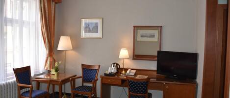 A hotel room with a bed, two chairs, a desk with a lamp, a television, and framed pictures on the wall.
