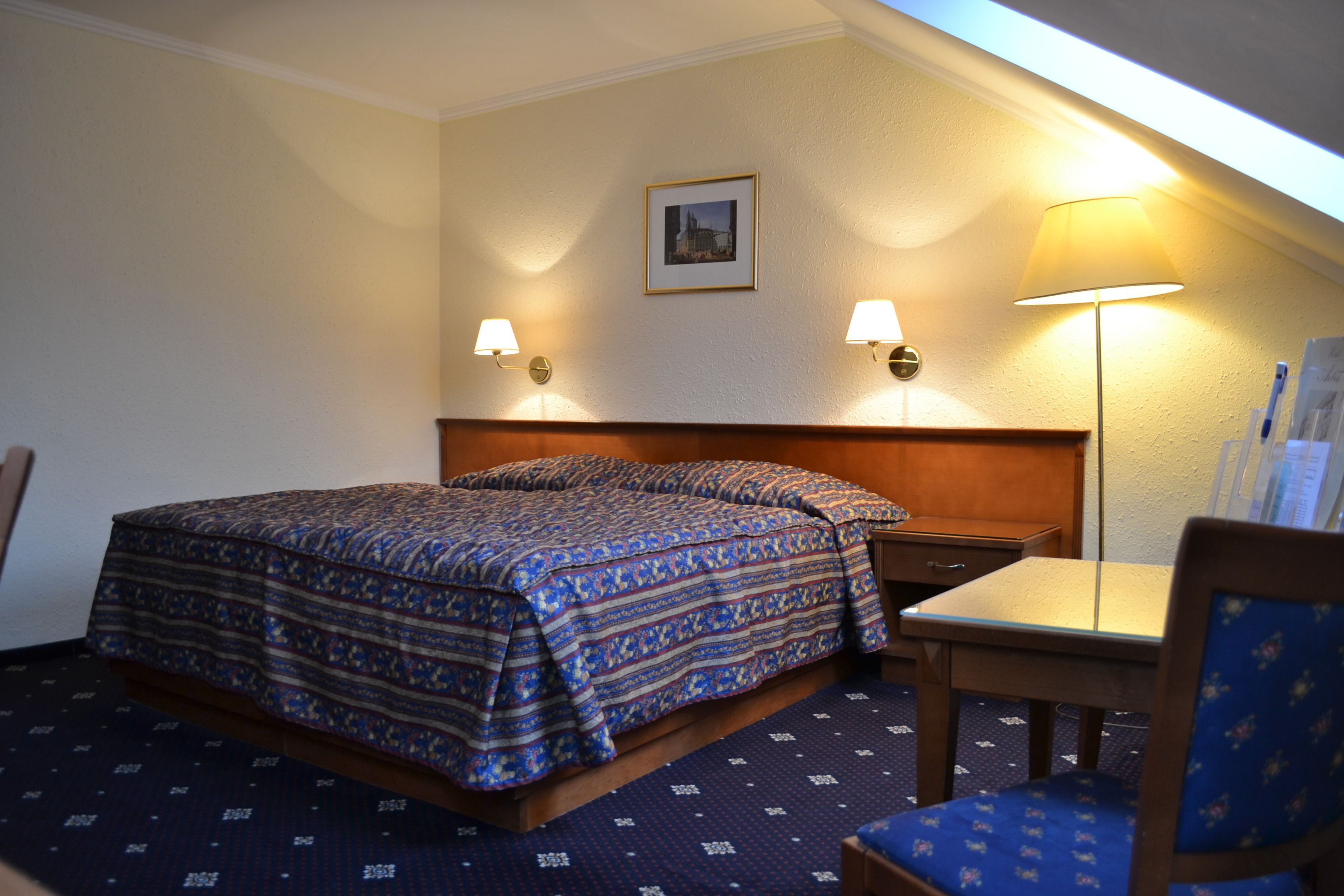 A hotel room with a bed, a desk, a chair, a lamp, and a picture on the wall.
