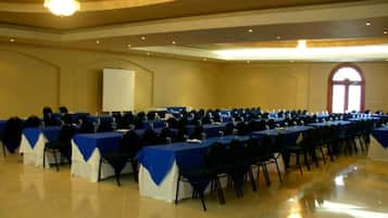 Un amplio salón de conferencias con filas de mesas y sillas, una pizarra y una pantalla de proyector.
