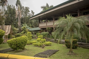 A building with a covered balcony, surrounded by well-maintained gardens and palm trees. - Hotel Suerre (Guapiles)