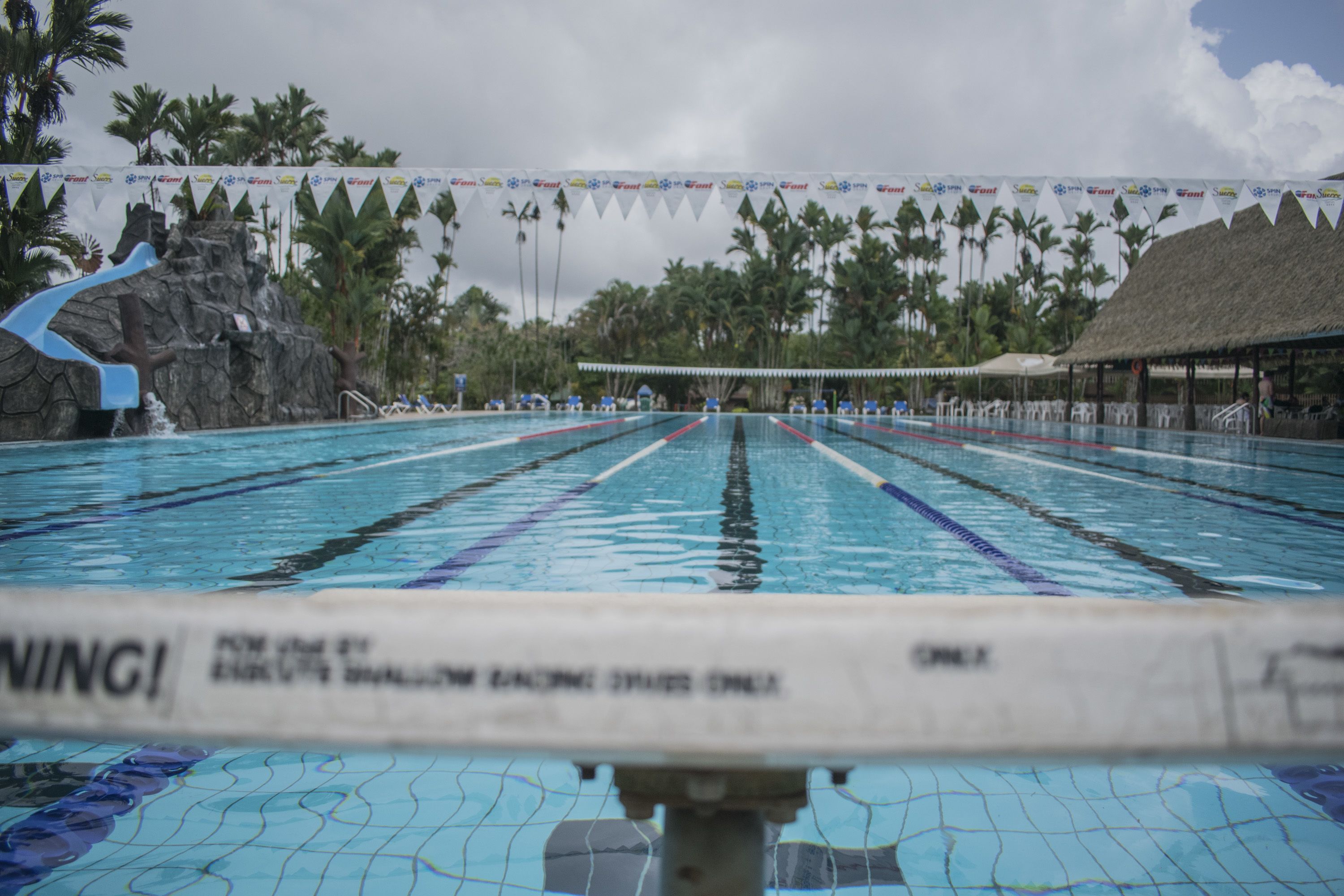 A swimming pool with lane dividers, a water slide, and palm trees in the background.