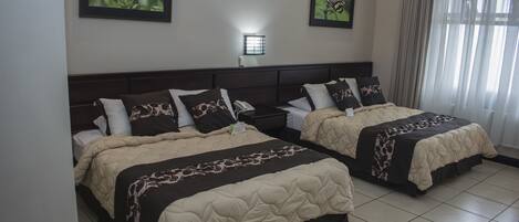 Habitación de hotel con dos camas, cada una con cabecera oscura y ropa de cama color beige. Hay dos cuadros en la pared, una lámpara de noche y una ventana con cortinas.