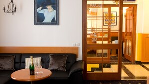 A room with a black leather sofa, a small wooden table with a bottle and glasses, a framed painting, and a wooden door leading to another room. - Hotel Máchova (Prague)