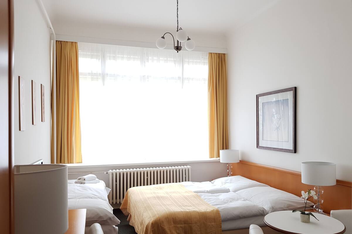 A hotel room with two beds, a desk, a chair, a round table with a lamp, a radiator, and a window with curtains.