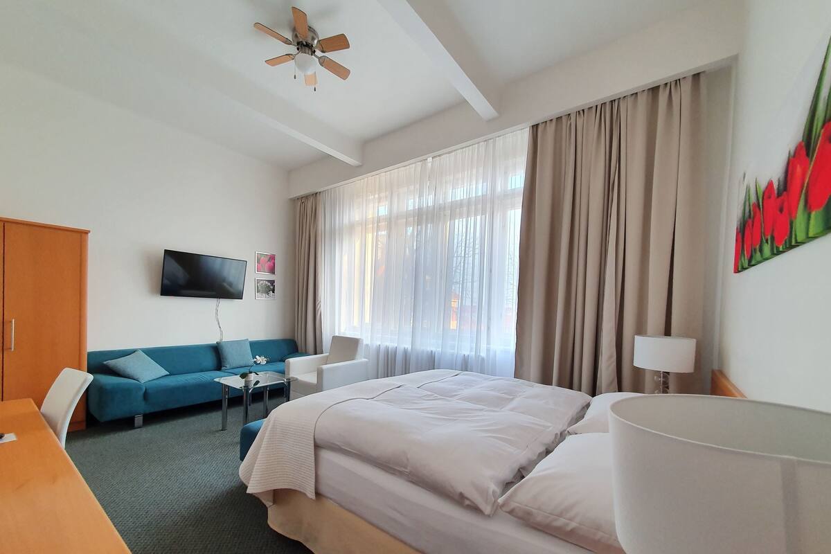 A hotel room with a bed, a blue sofa, a white armchair, a TV on the wall, and a ceiling fan.