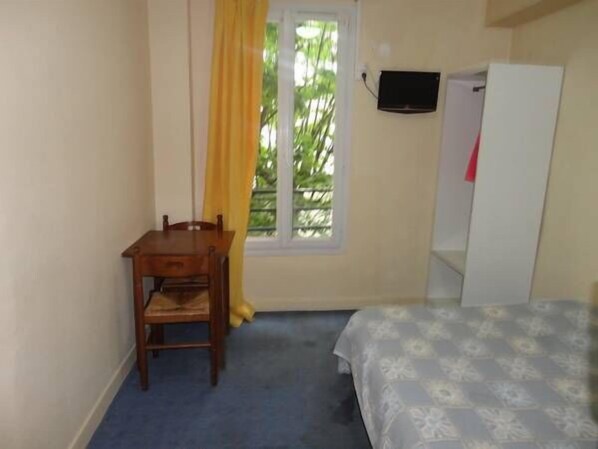 A small room with a bed, a desk, and a chair. There is a window with yellow curtains and a television mounted on the wall.