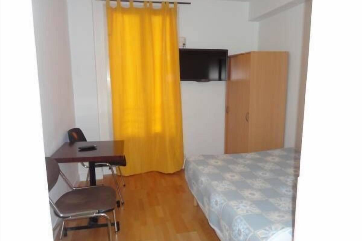 A small room with a bed, a desk, a chair, a wardrobe, and a TV on the wall.