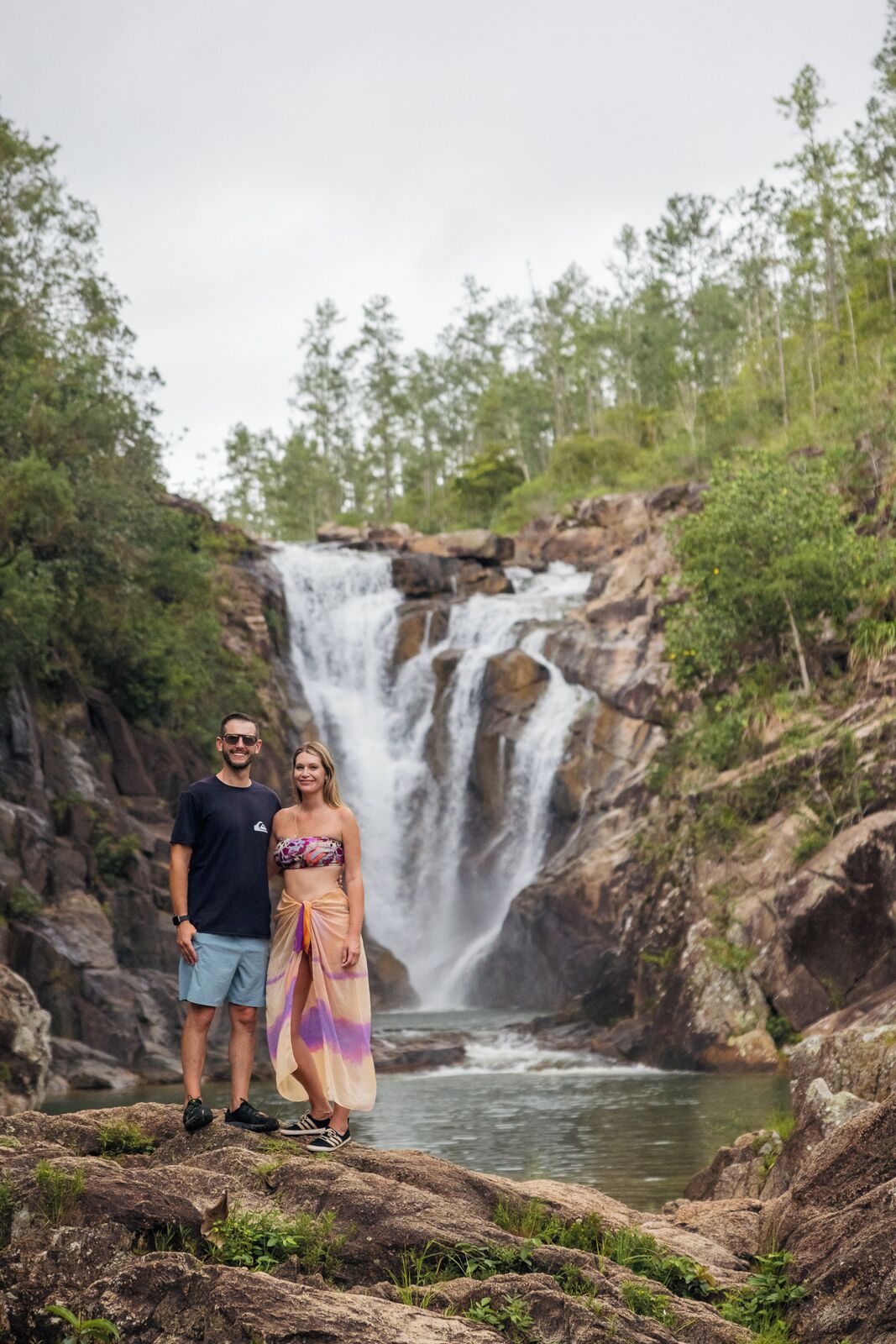Una pareja de pie sobre rocas, con una cascada al fondo.