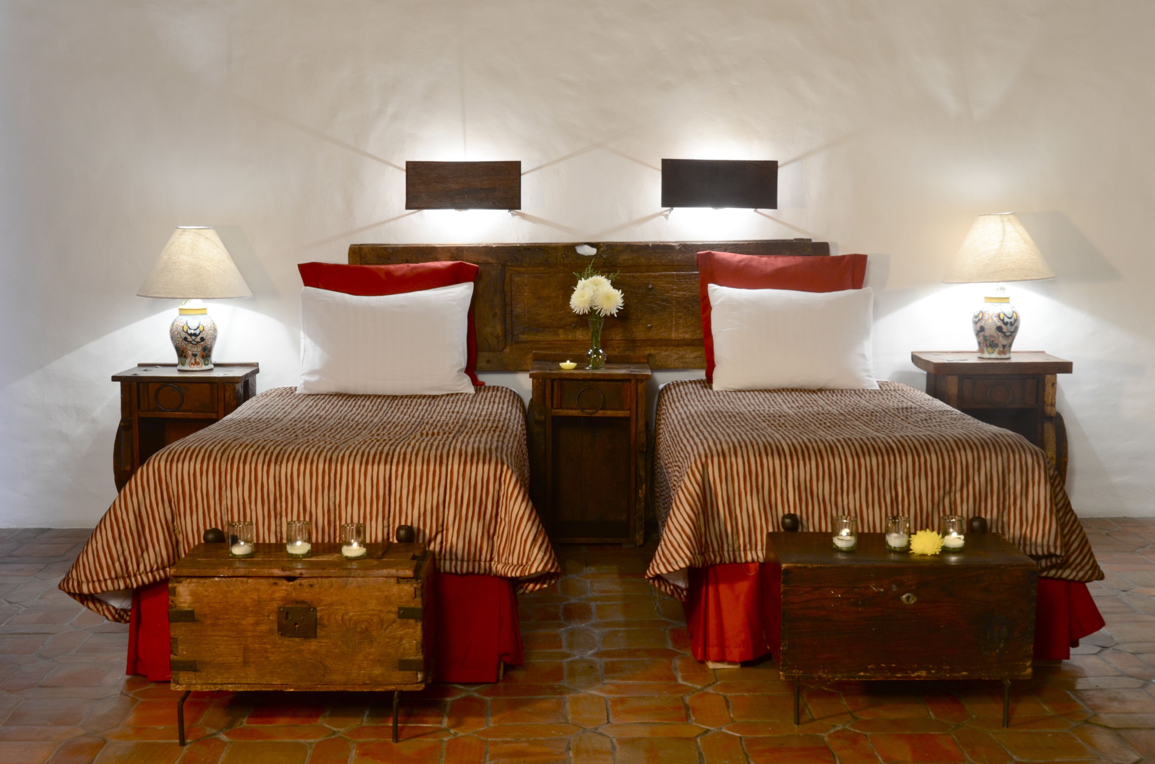 A room with two beds, each with a striped bedspread and red pillows, wooden nightstands with lamps, and a wooden headboard with a vase of flowers.