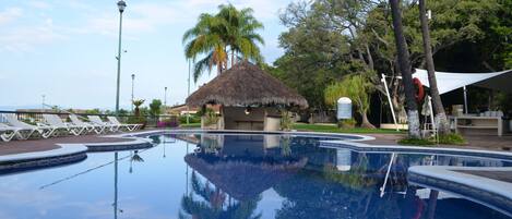 Una piscina con sillas de descanso, una cabaña con techo de paja y palmeras.