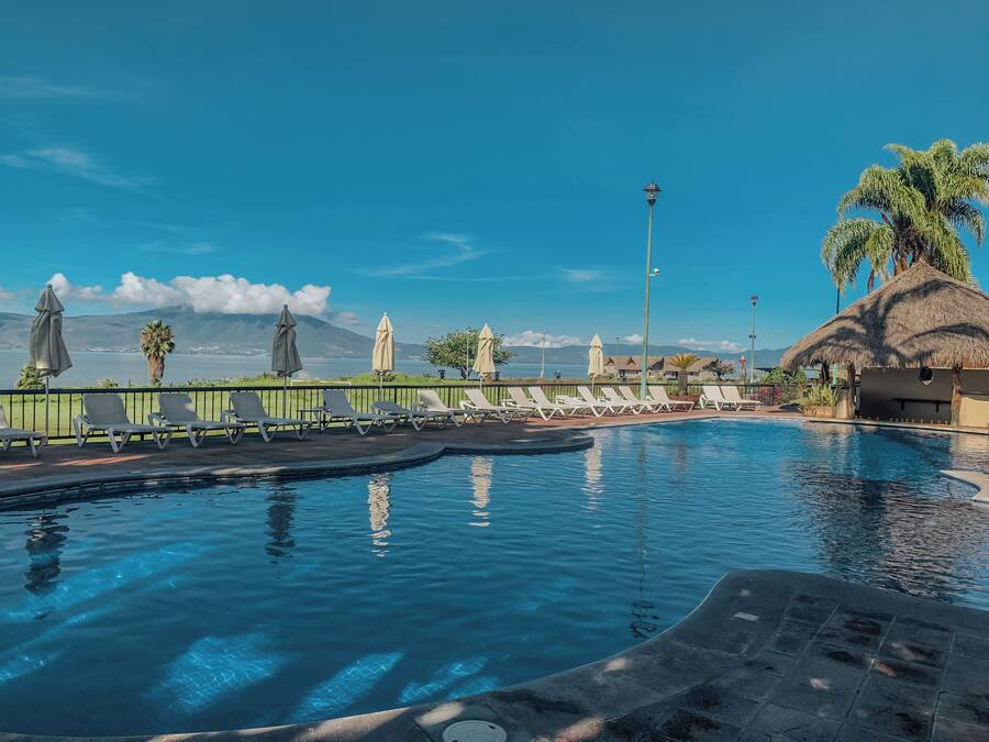 Una piscina con sillas de descanso y sombrillas, una estructura de techo de paja, palmeras y una vista de montañas al fondo.