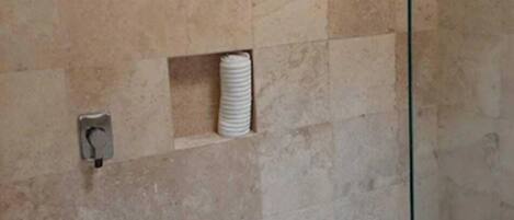 A shower area with beige tiles, a glass door, and a small shelf with a white towel.