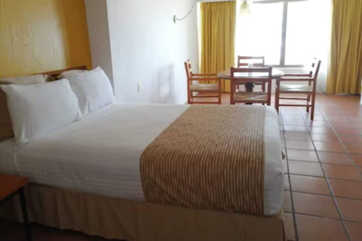A hotel room with a large bed, a wooden table, and chairs, and a view of the outdoors through the window.