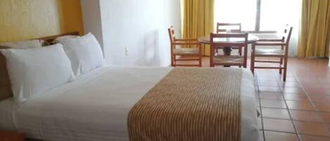 A hotel room with a large bed, a wooden table, and chairs, and a view of the outdoors through the window.