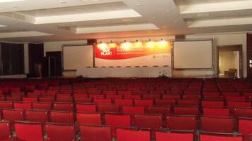 Un salón de conferencias con hileras de sillas rojas, un escenario con un cartel y una pantalla grande.