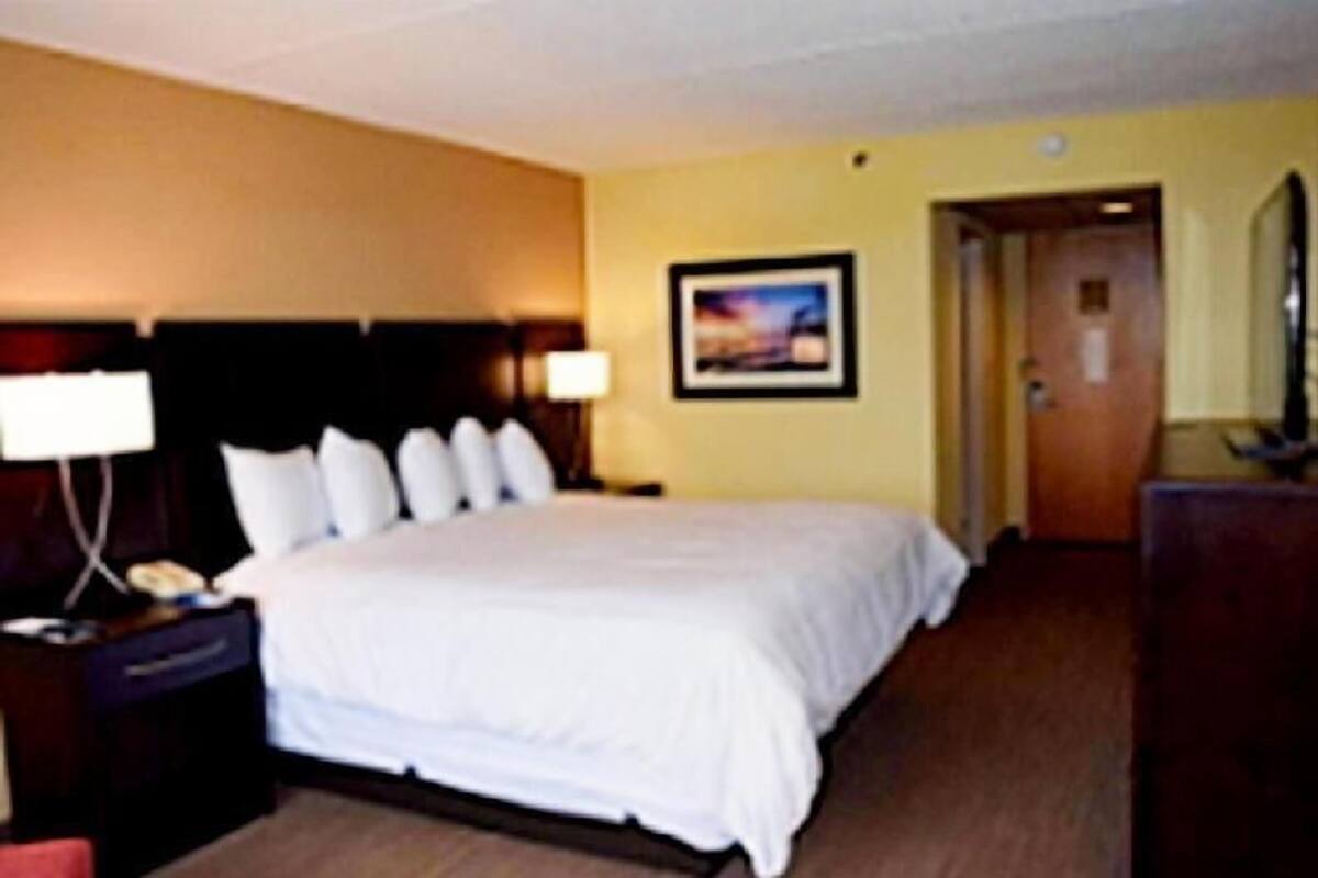 Quarto de hotel com uma cama, mesa de cabeceira, abajur, quadro na parede e porta que dá para outro cômodo.