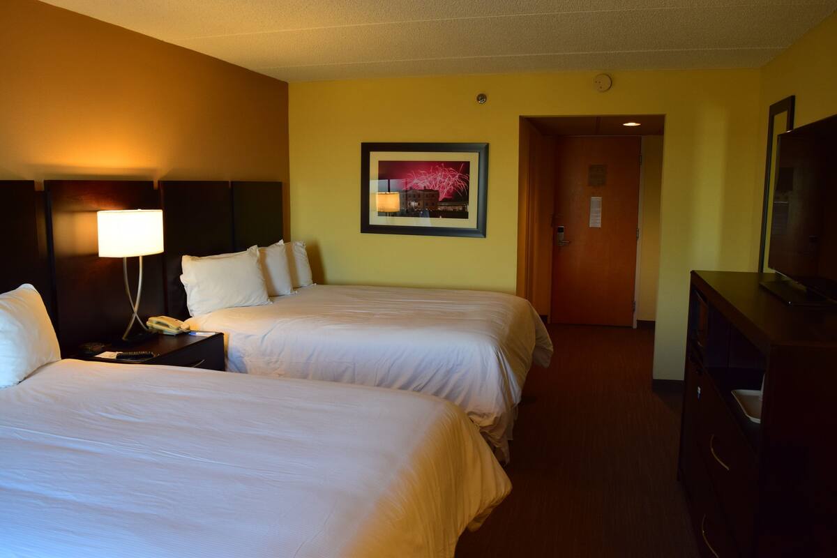 Quarto de hotel com duas camas, uma escrivaninha com luminária, televisão e um quadro na parede.