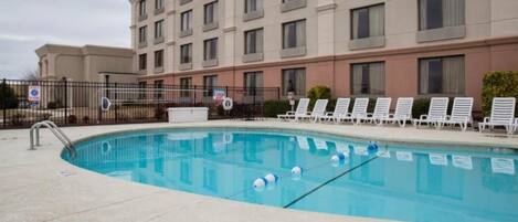 Uma piscina com espreguiçadeiras, cercas e um edifício ao fundo.