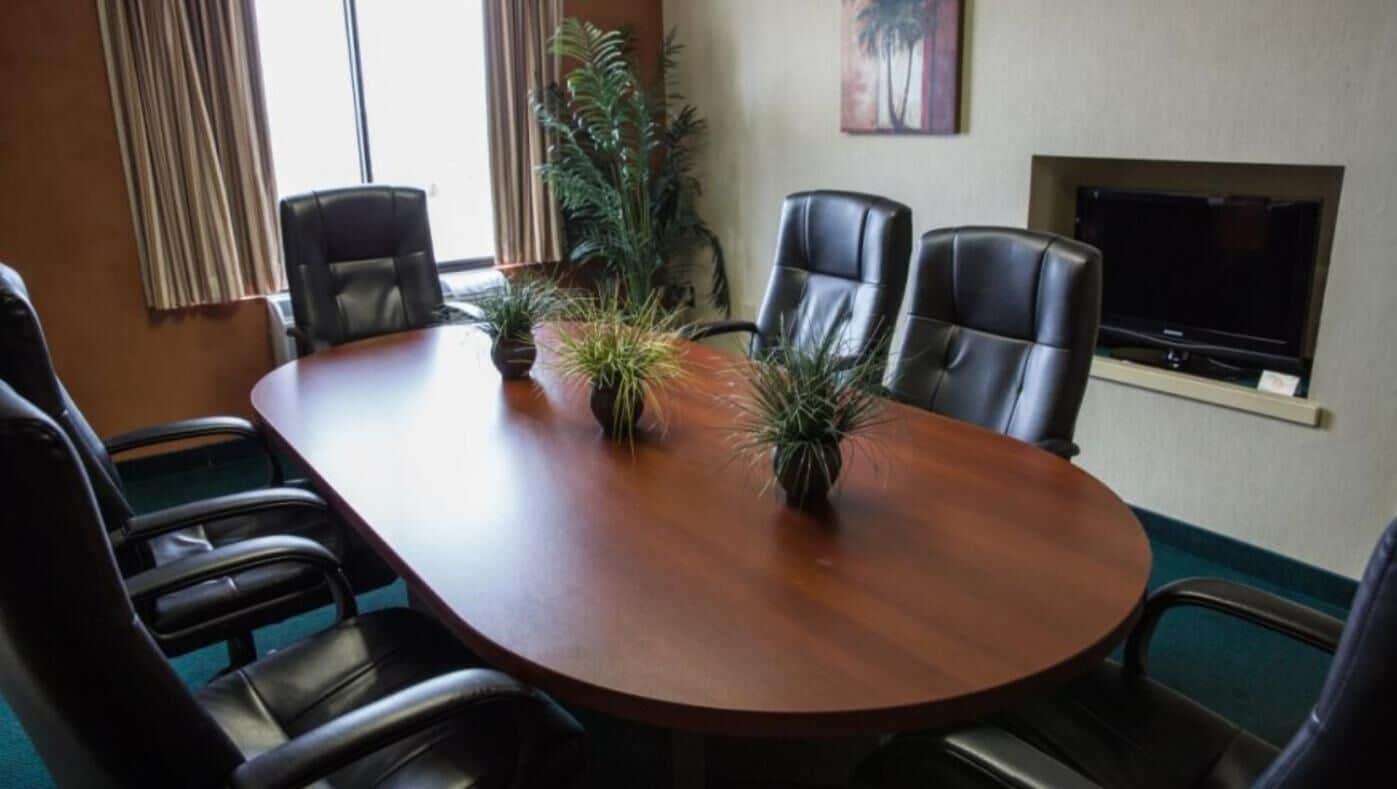 A conference room with a wooden table, four leather chairs, two potted plants, a television, and a painting on the wall.