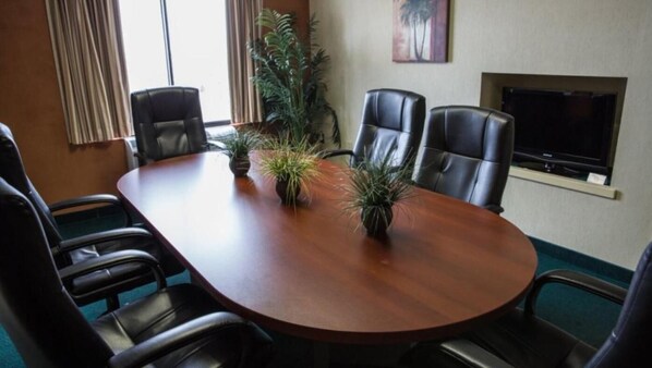 A conference room with a wooden table, four leather chairs, two potted plants, a television, and a painting on the wall.