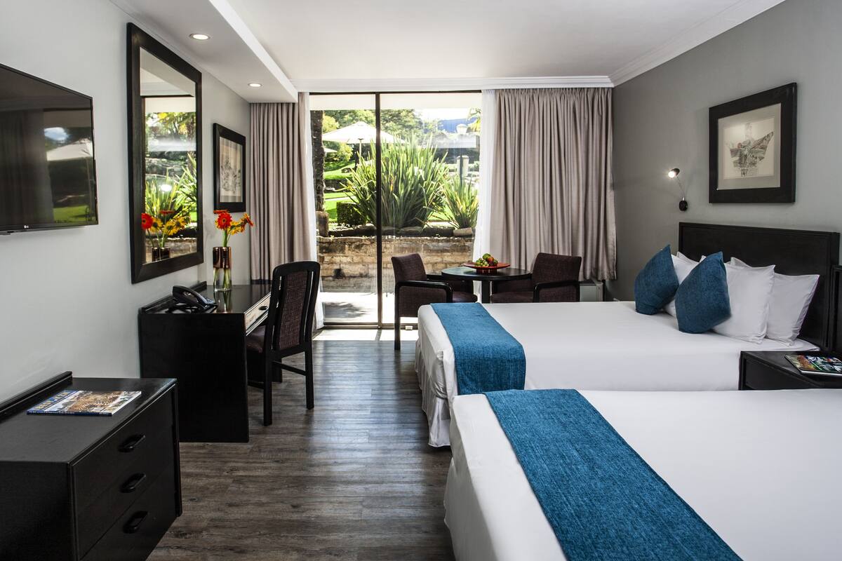 A hotel room with two beds, a desk, a chair, a TV, and a view of the garden.