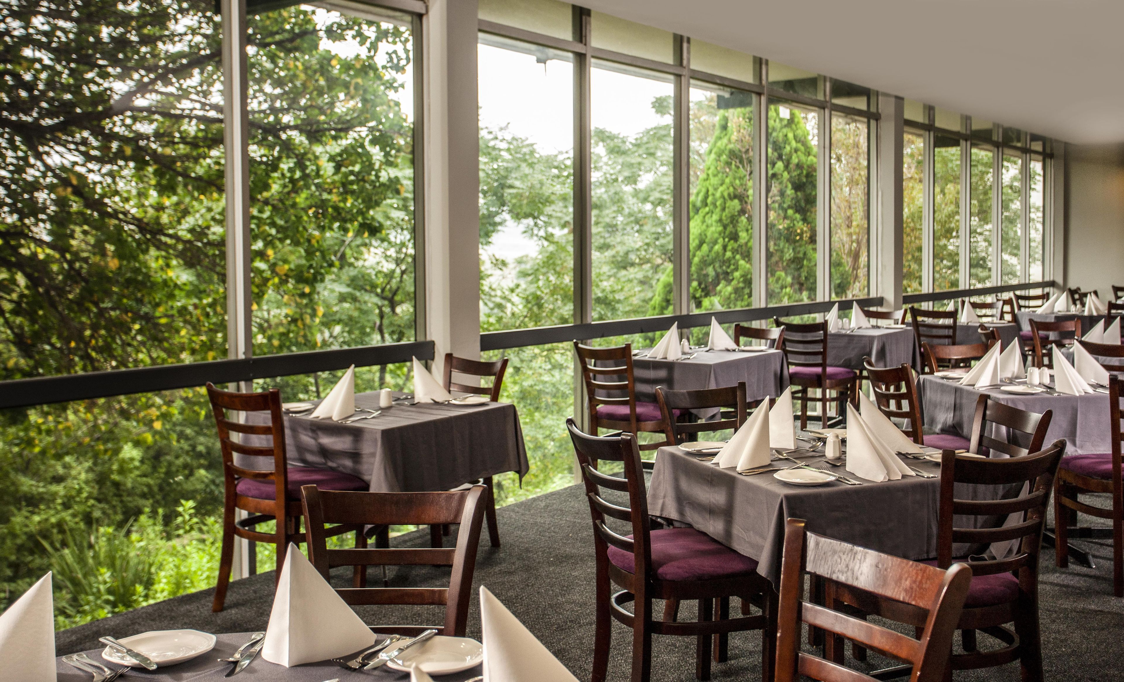 Un restaurant avec des tables dressées pour le service, de grandes fenêtres et une vue sur les arbres.