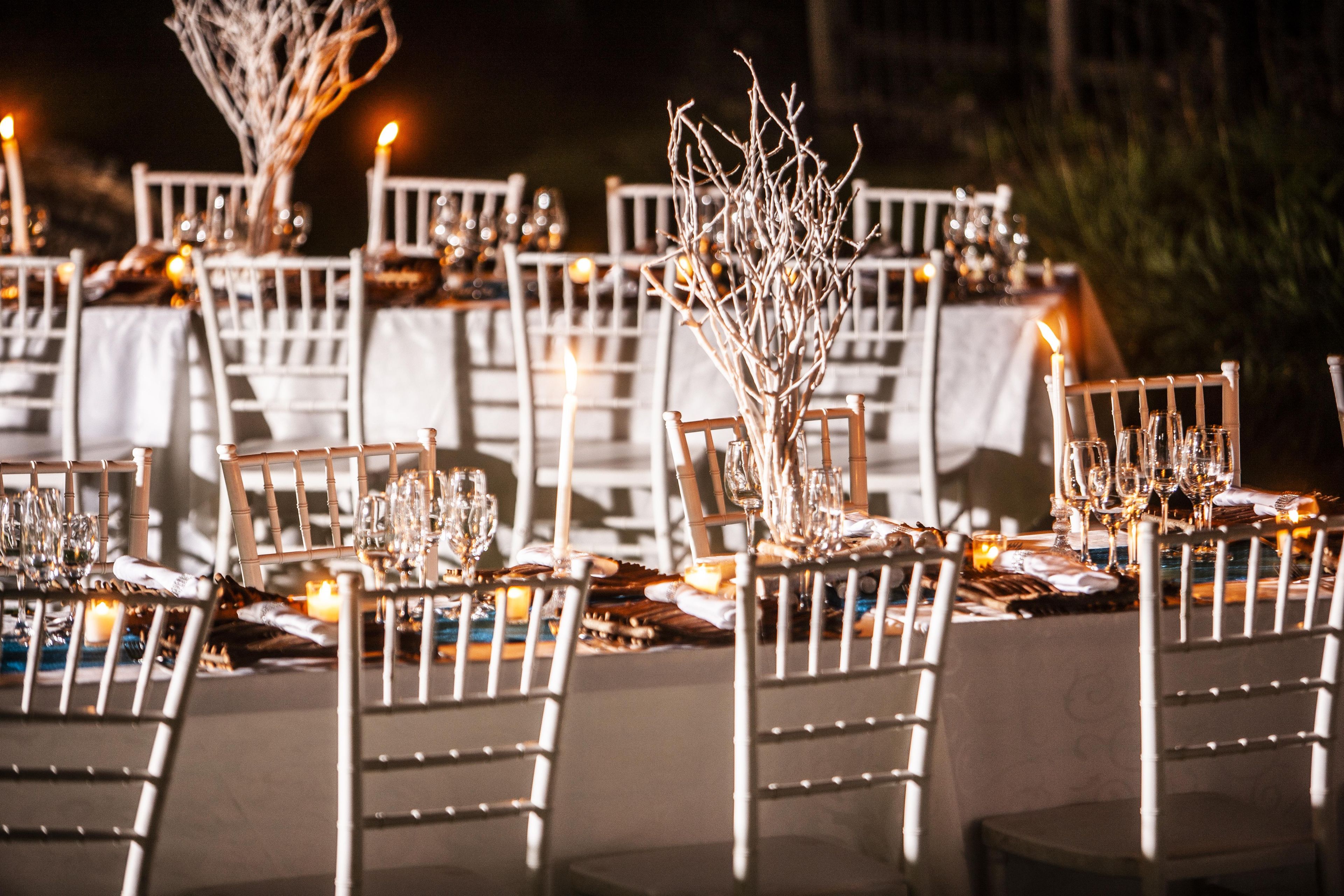 A formal outdoor dining setup with tables set for multiple guests, featuring white chairs and candlelight.