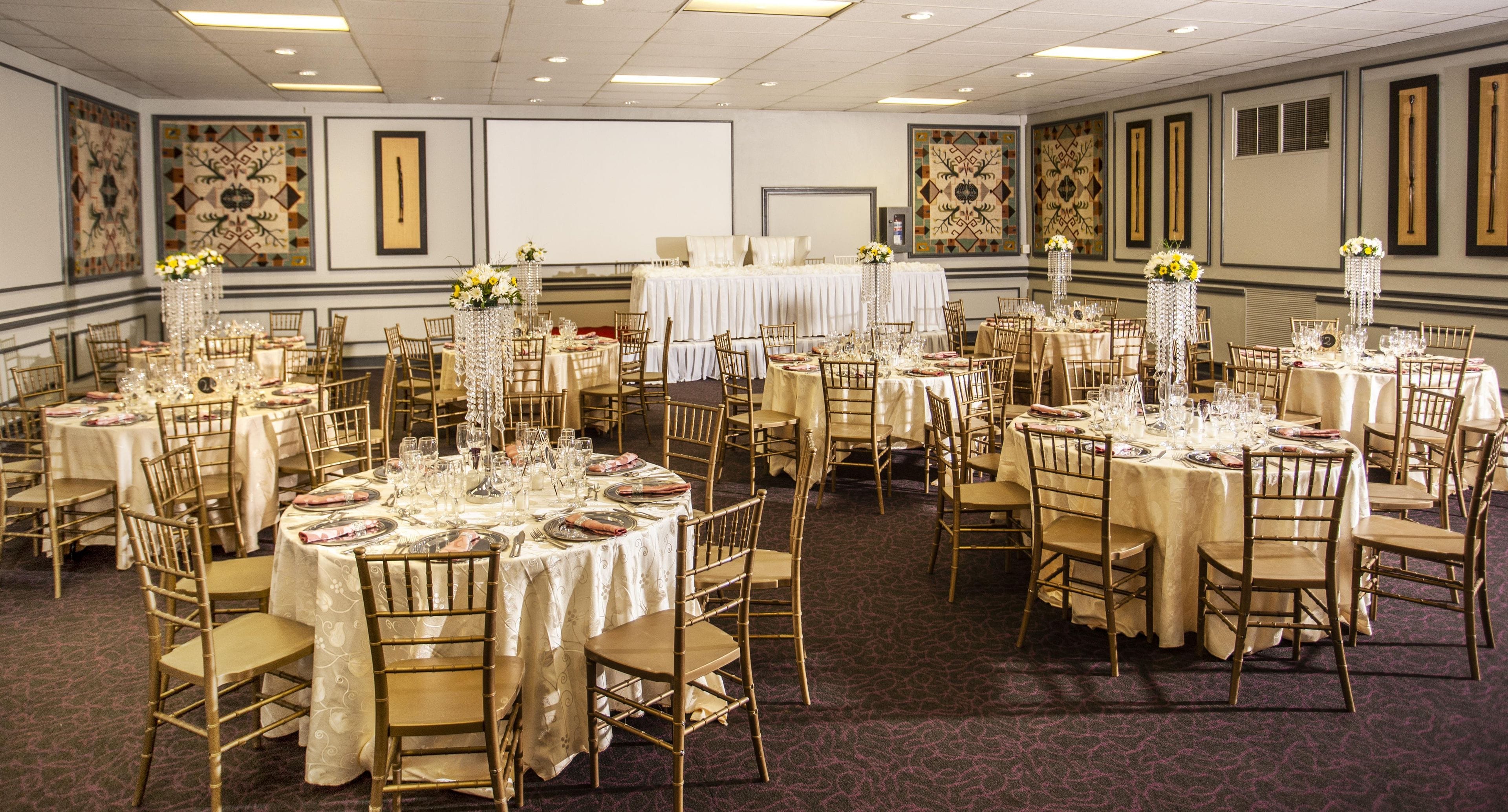Een feestzaal met ronde tafels, stoelen en bloemstukken als decoratie.
