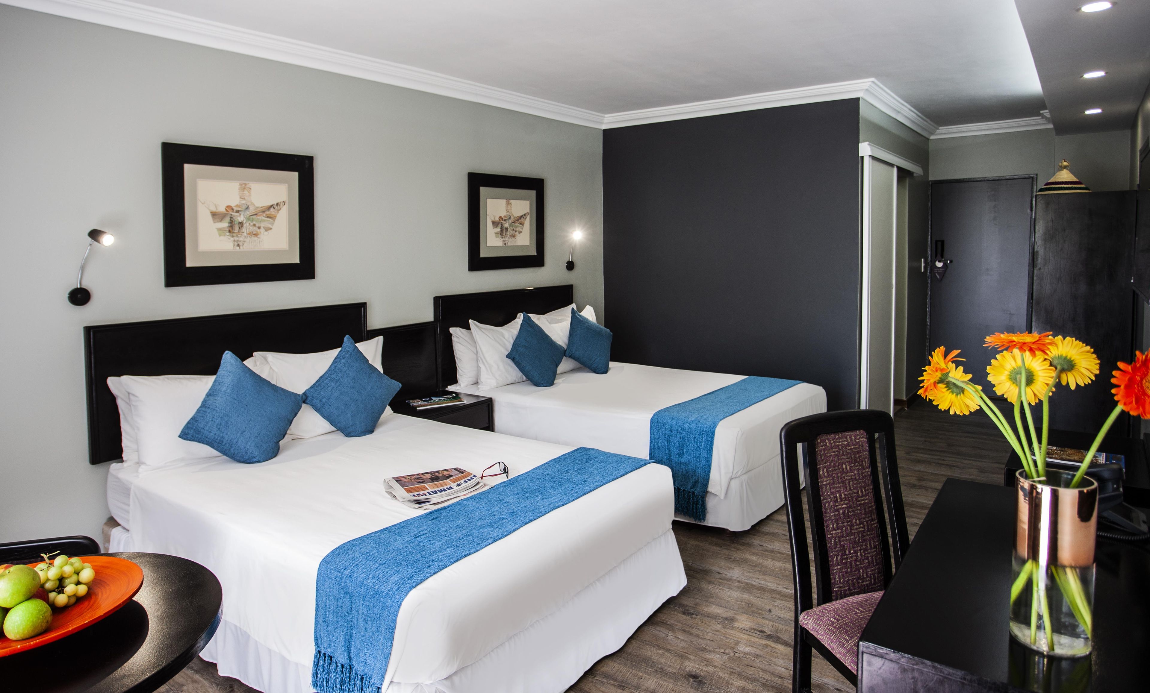 Een hotelkamer met twee bedden, een eettafel, een vaas met bloemen en ingekaderde foto's aan de muur.