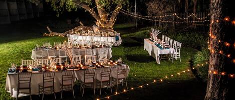 Un área de comedor al aire libre con mesas dispuestas para una comida, rodeada de árboles y luces colgantes.