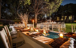 Un área de comedor al aire libre con mesas dispuestas para una comida, rodeada de árboles y luces colgantes.