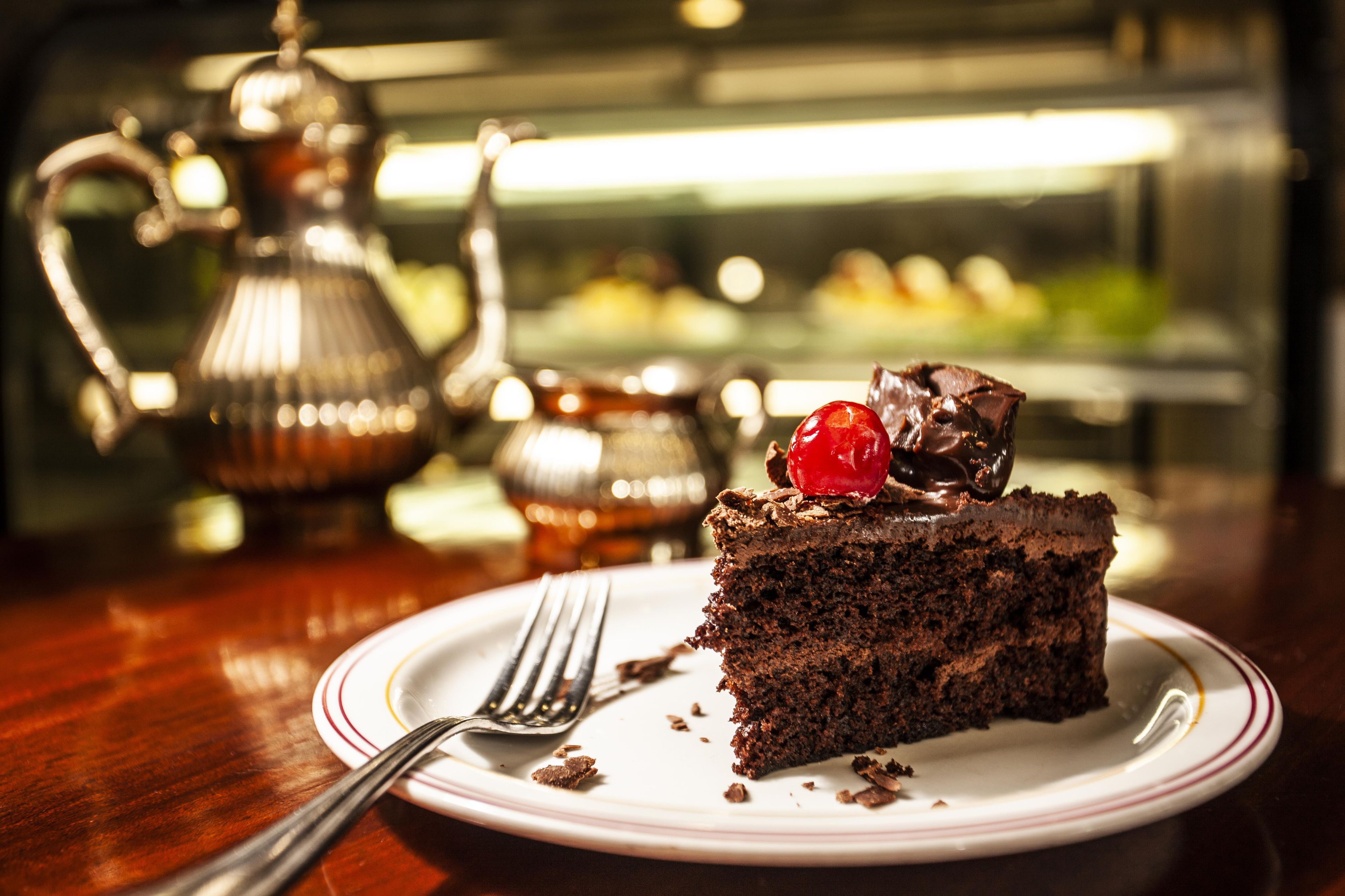 A slice of chocolate cake with a cherry on a white plate with a fork beside it.
