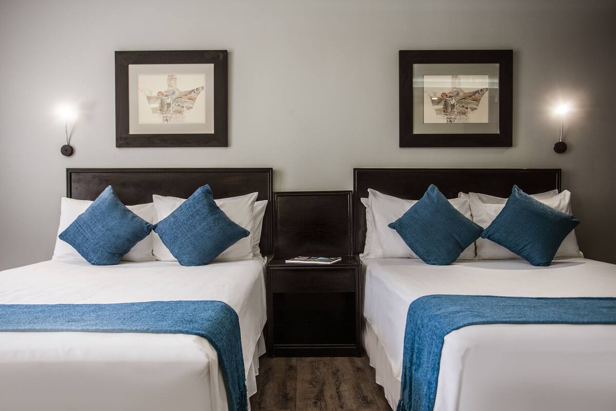 A hotel room with two beds, each with white linens and blue pillows, a nightstand with a book, and framed artwork on the wall.
