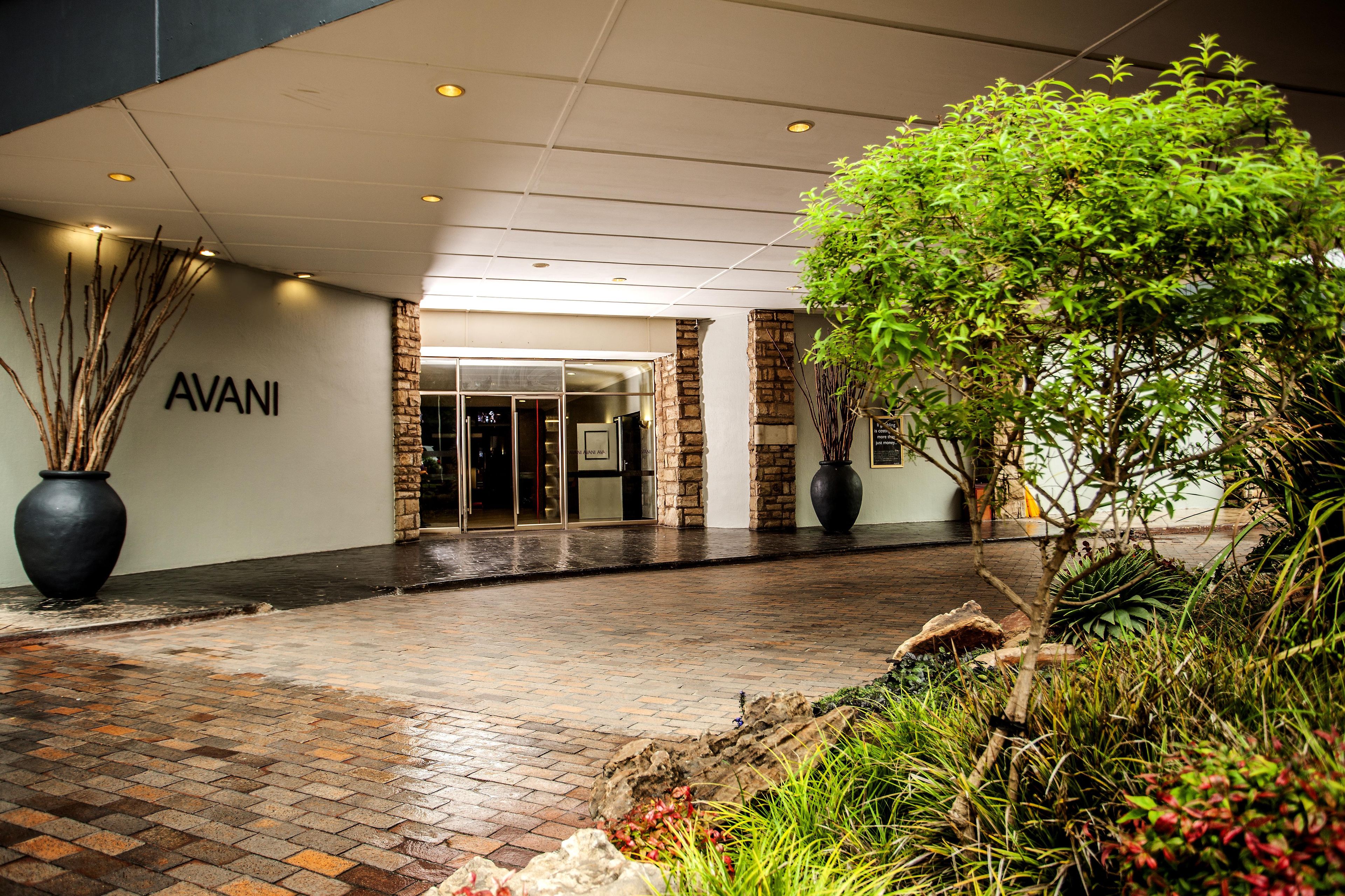 A hotel entrance with a brick pathway leading to a glass door entrance, surrounded by potted plants and a stone wall.