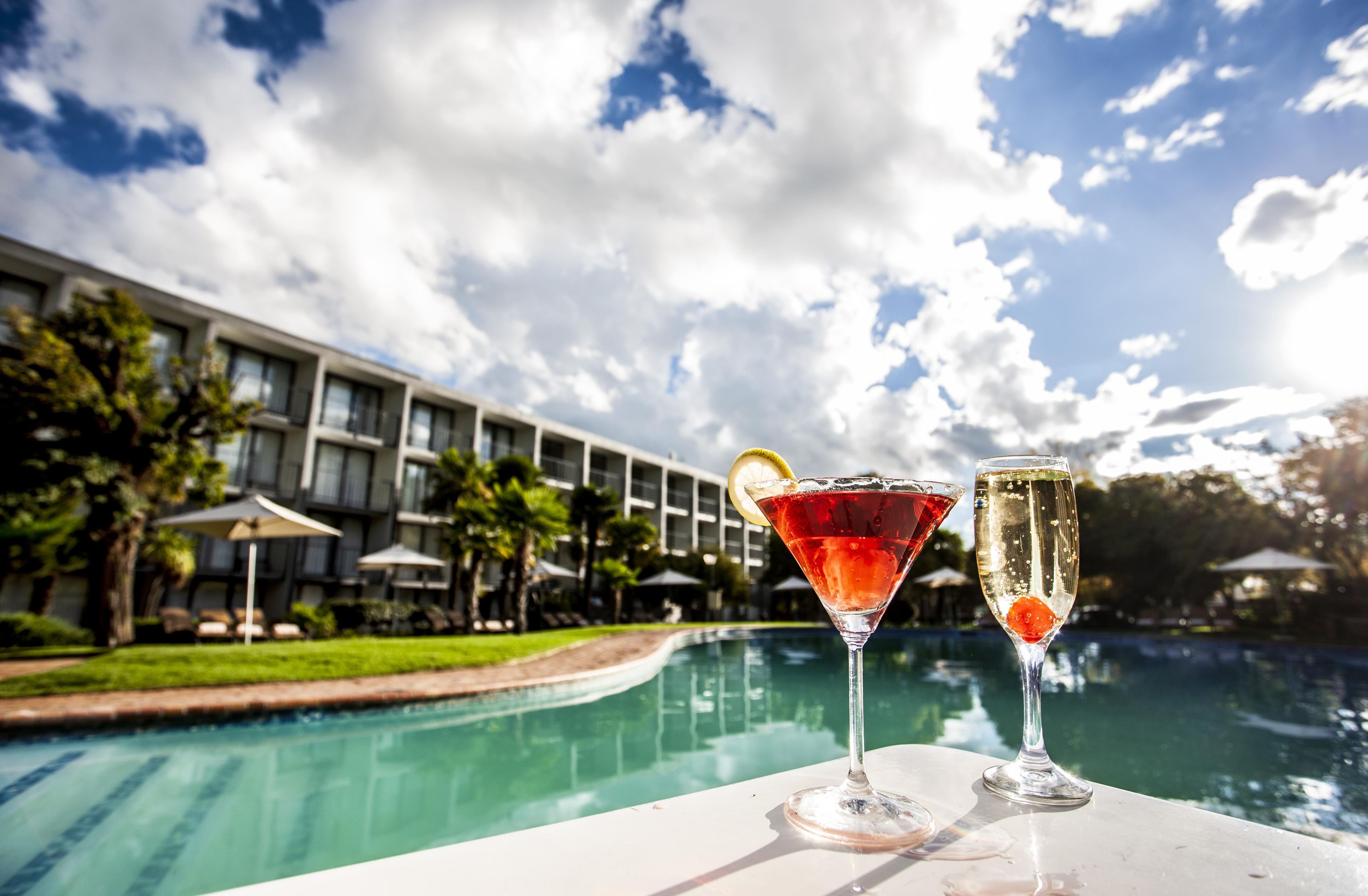 A poolside view with two cocktails, a building with a modern design, and a clear sky.