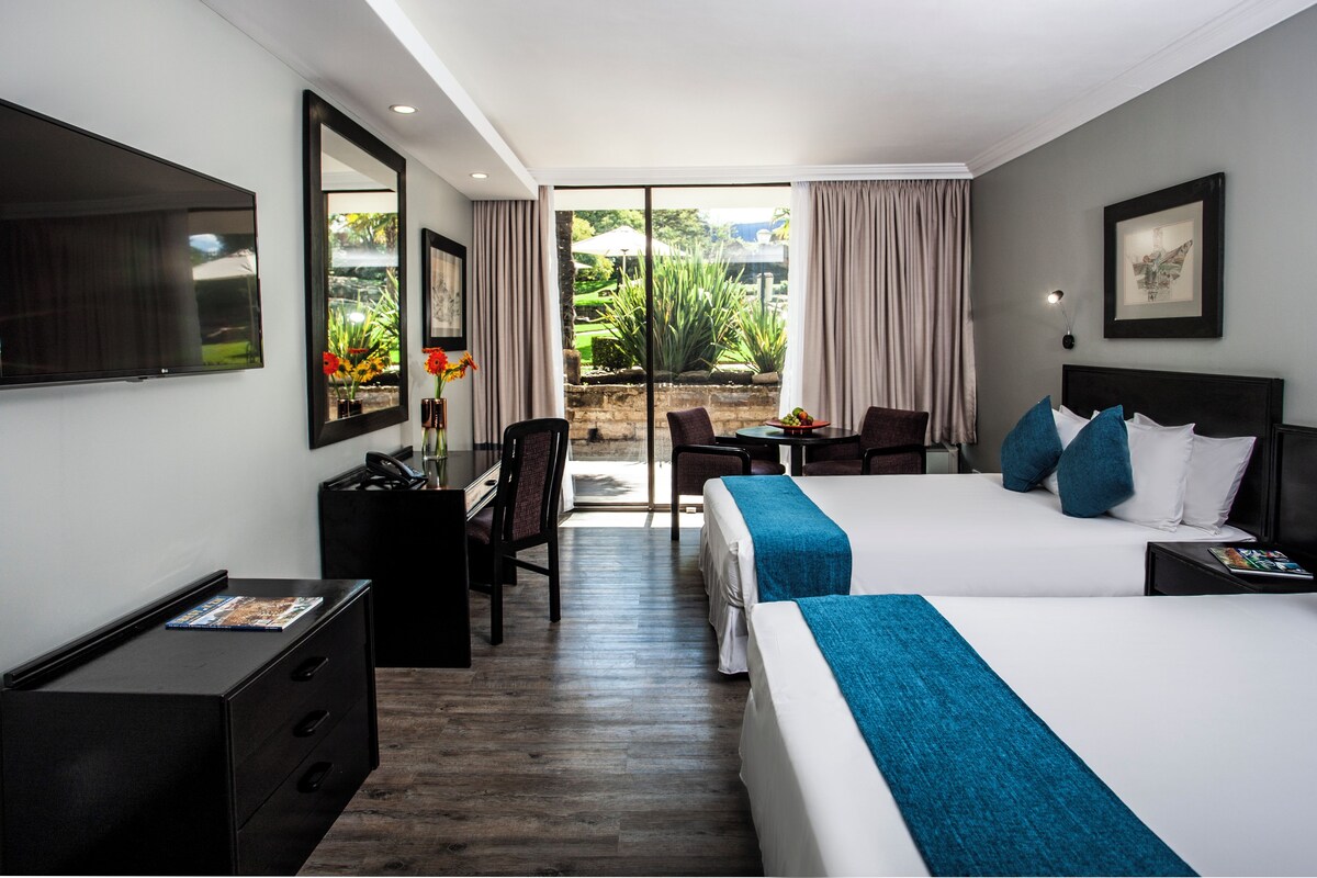 A hotel room with a large bed, a desk with a computer, a TV, and a view of the garden.